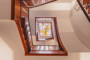 Spiral wooden staircase looking up towards a colorful stained glass skylight at Address_16.