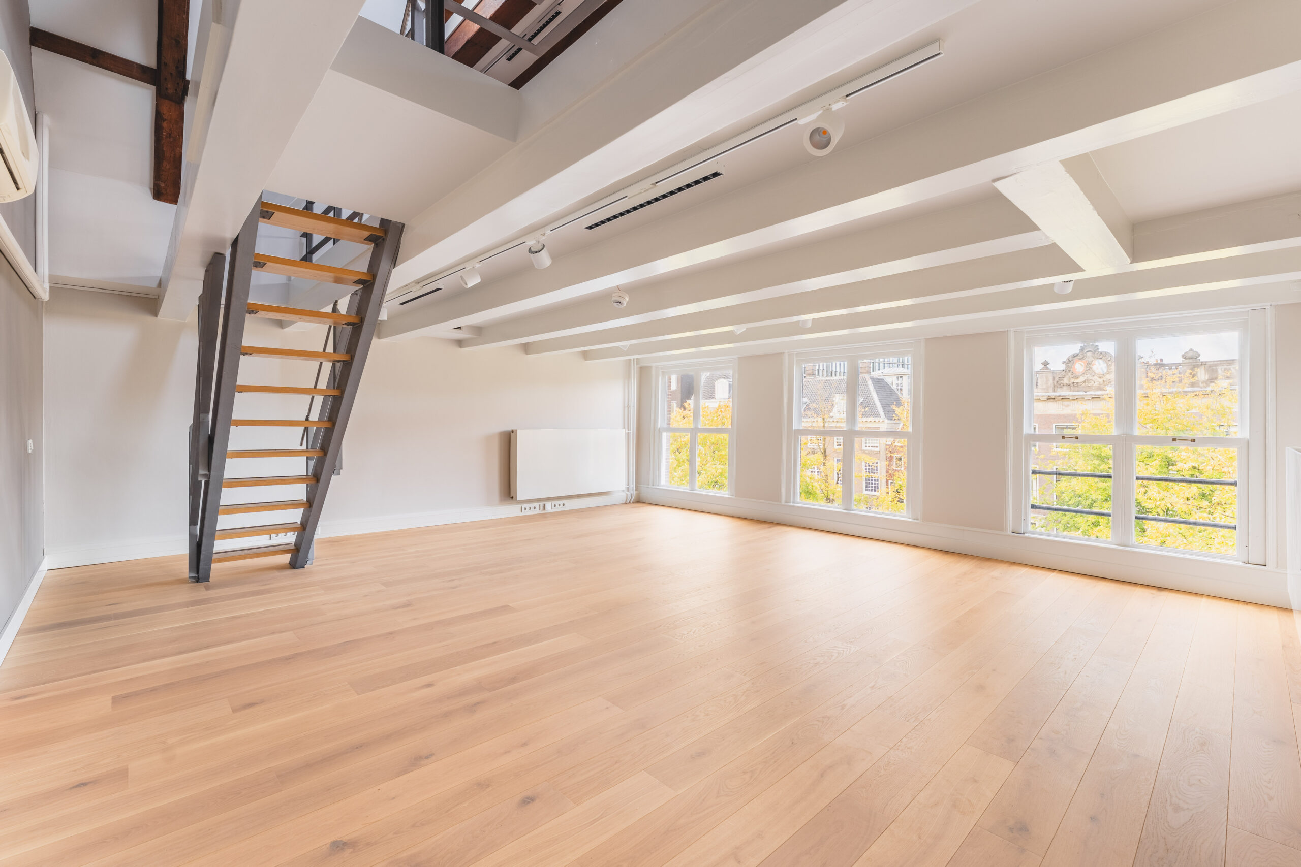 Bright loft-style room with wooden flooring, large windows, and a modern open staircase.