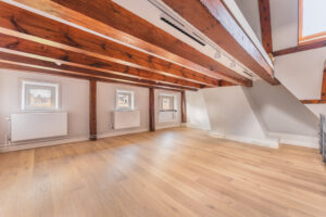 Bright attic room with wooden beams, light oak flooring, and small windows along one wall at Address_19.
