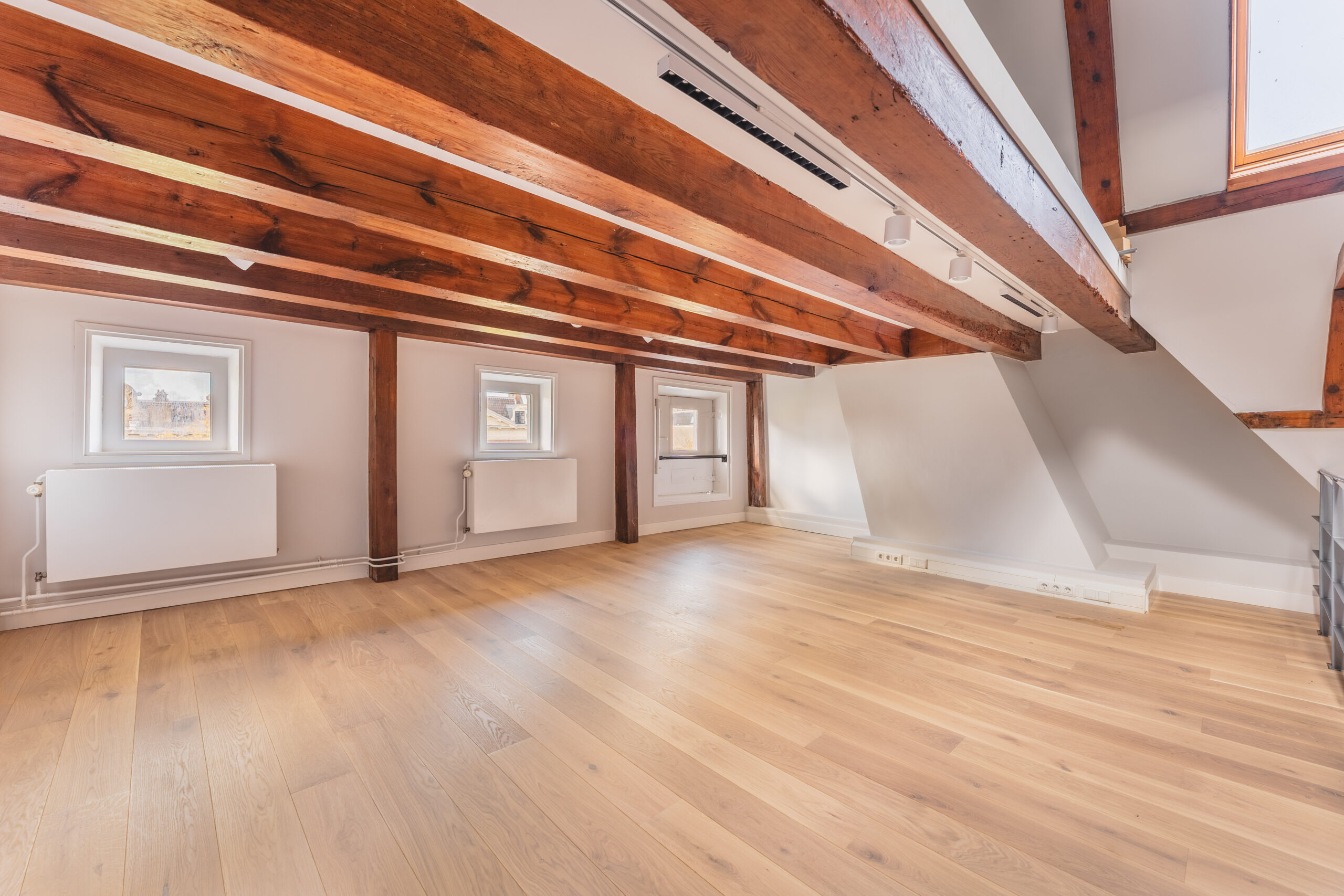 Bright attic room with wooden beams, light oak flooring, and small windows along one wall at Address_19.