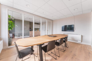 Modern meeting room with a wooden conference table, black chairs, wall-mounted TV, and glass partition revealing a kitchenette.