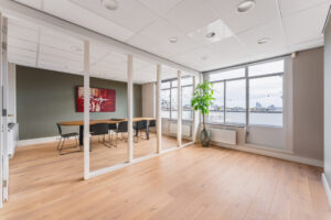Modern office space with a glass partition, wooden flooring, large windows with a city view, and a minimalist meeting area.