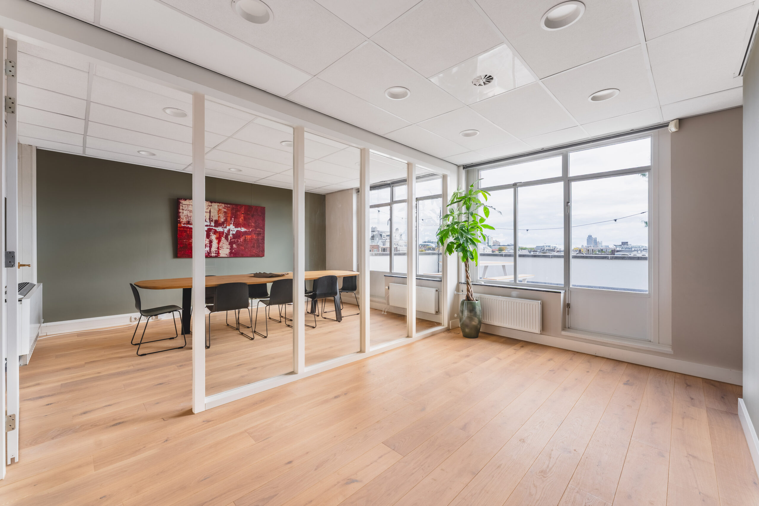 Modern office space with a glass partition, wooden flooring, large windows with a city view, and a minimalist meeting area.