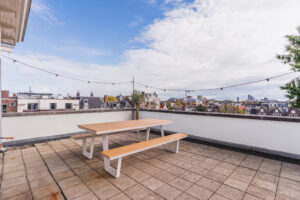 Rooftop terrace with a wooden picnic table, string lights, and a view over city rooftops.