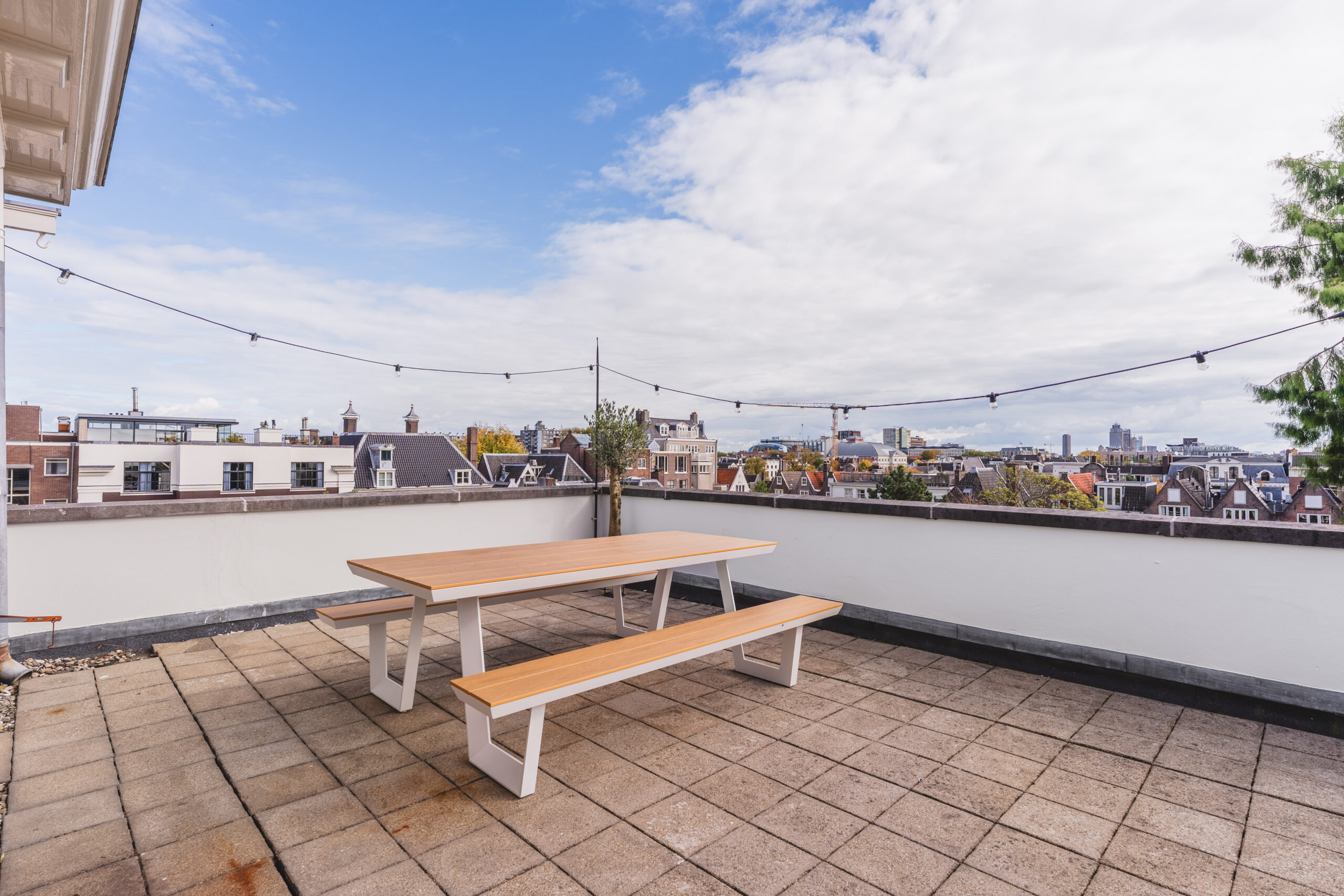 Rooftop terrace with a wooden picnic table, string lights, and a view over city rooftops.