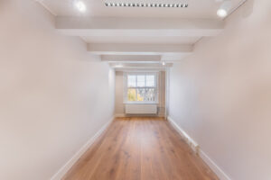 Bright, narrow room with wooden flooring, white walls, exposed ceiling beams, and a large window overlooking a cityscape.