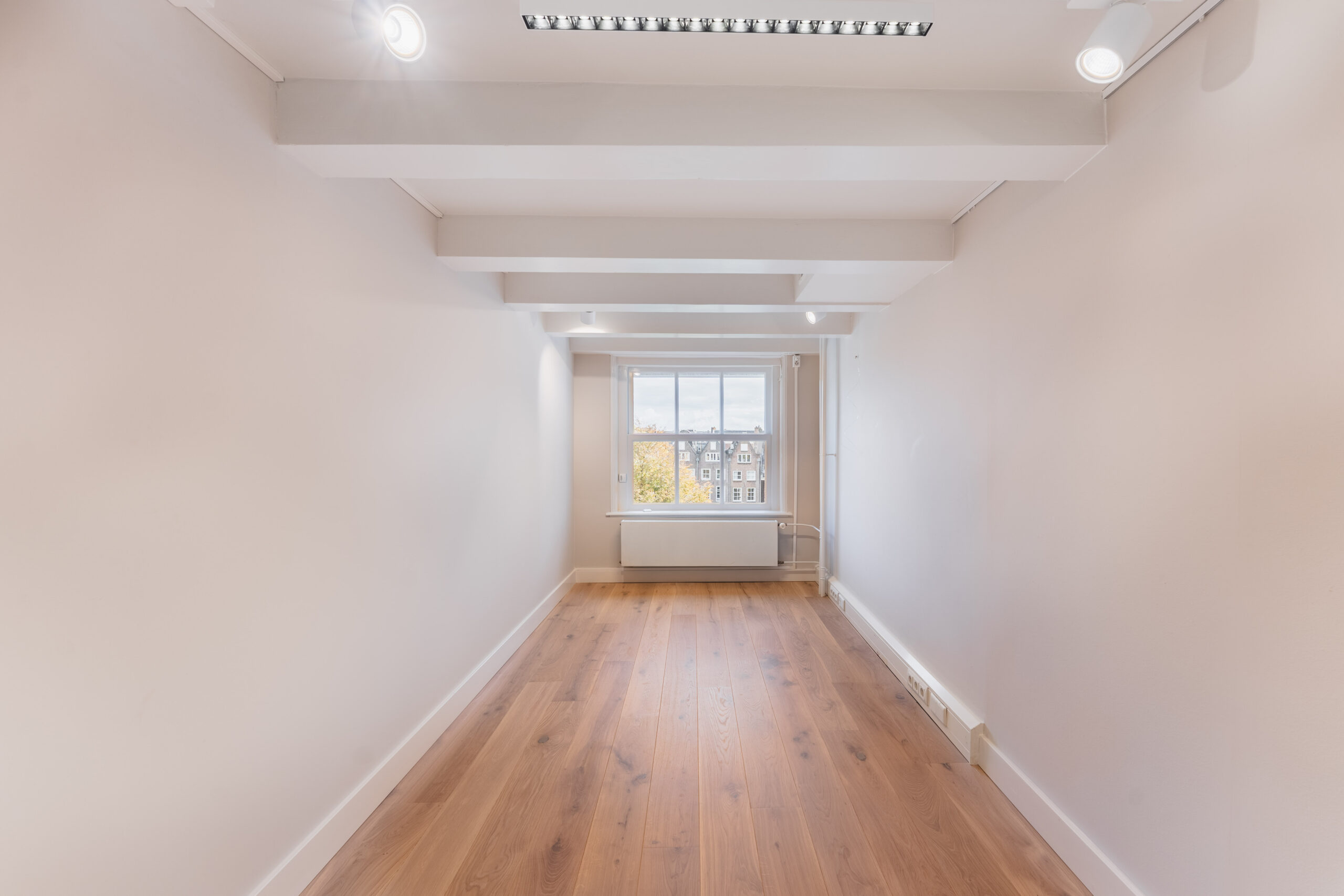 Bright, narrow room with wooden flooring, white walls, exposed ceiling beams, and a large window overlooking a cityscape.
