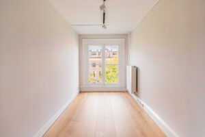 Minimalist, narrow room with light wooden flooring, white walls, and a large window overlooking classic architecture.