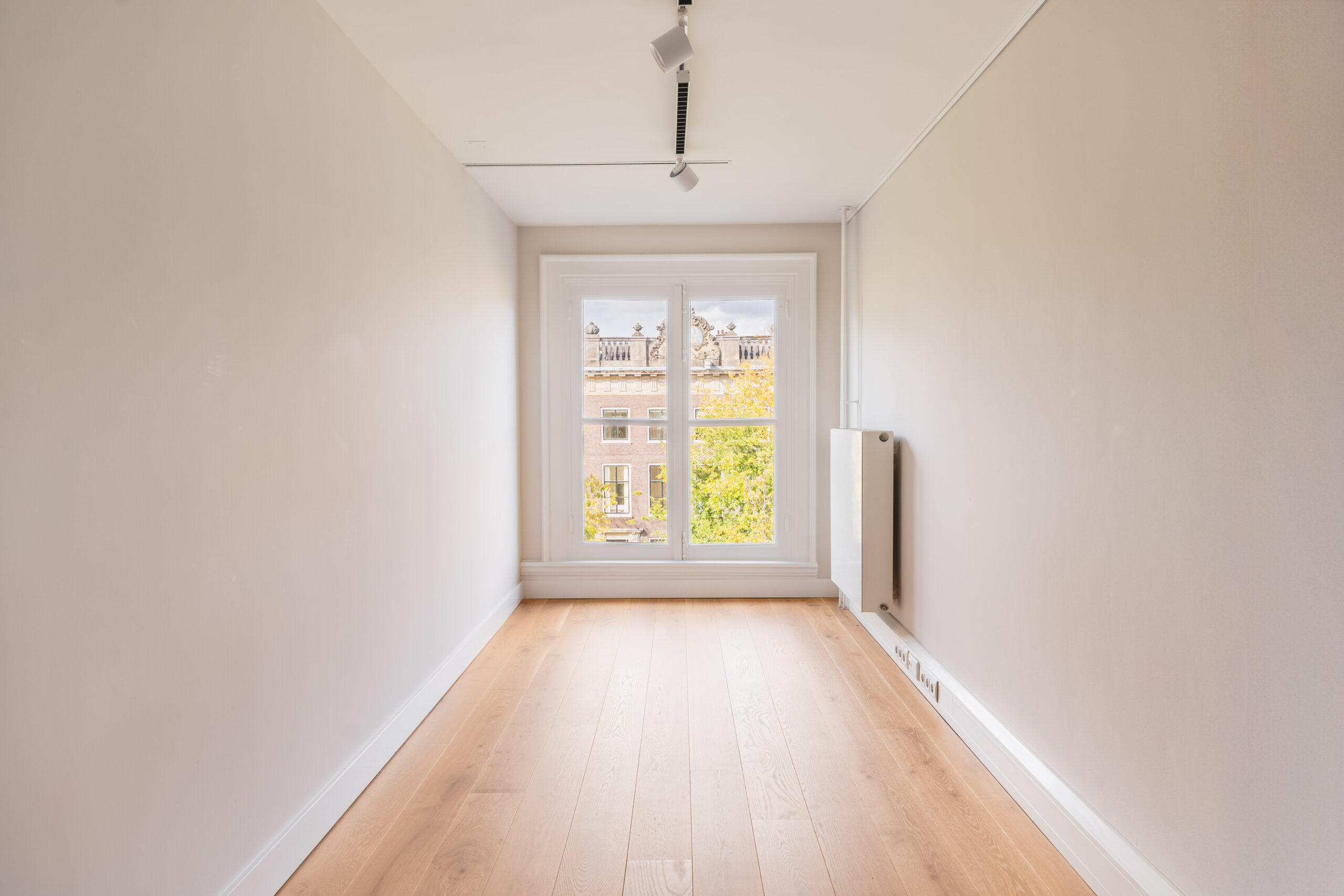Minimalist, narrow room with light wooden flooring, white walls, and a large window overlooking classic architecture.