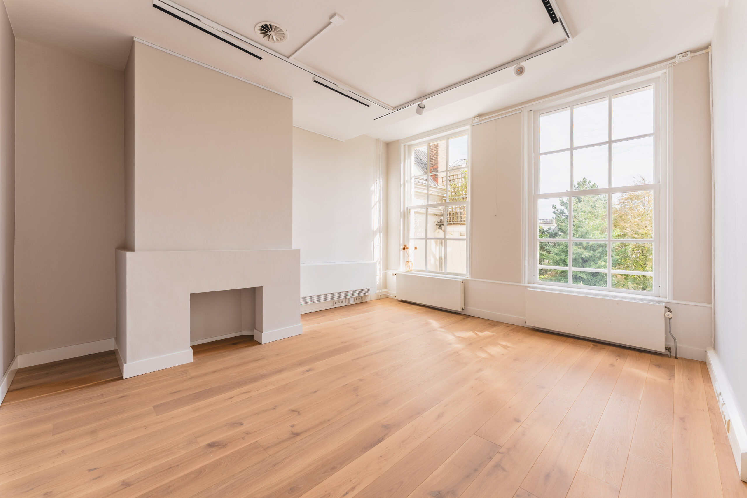 Bright, empty room at Adress_9 with large windows, light wooden floor, and a minimalist fireplace.