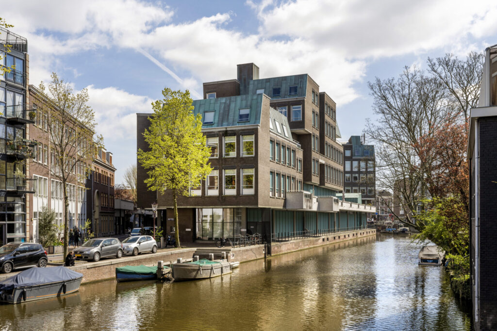Kantoorgebouw aan een Amsterdamse gracht met aangemeerde boten en bomen in lentebladeren.