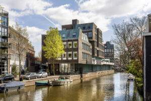 Modern canal-side buildings including the DSC_0621 office complex in a sunny Amsterdam street view with boats and trees.