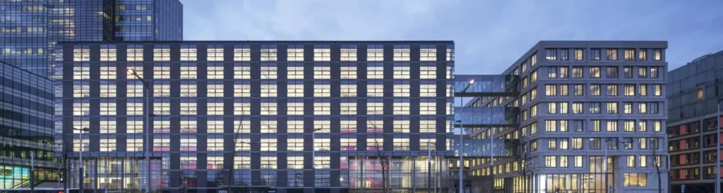 Voorgevel van het Piet Hein Building bij schemering, met verlichte ramen en moderne architectuur.