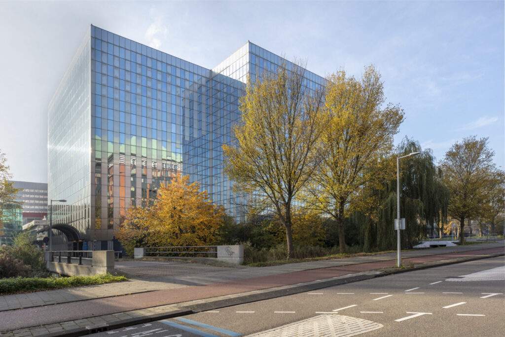 Glazen kantoorgebouw van MRP Flex Offices 13 weerspiegeld in herfstige omgeving met bomen en lege straat op de voorgrond.