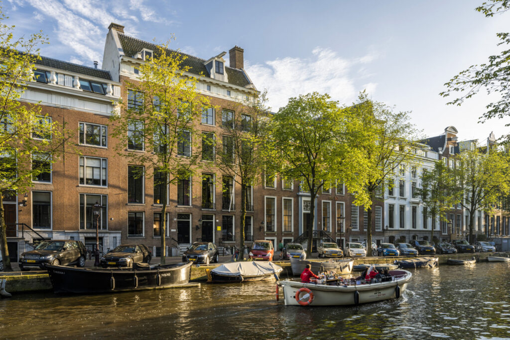 Zicht op de Herengracht in Amsterdam met historische grachtenpanden, geparkeerde auto's en een varende boot in het zonlicht.