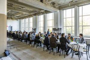 Groep mensen aan lange tafels in een industriële ruimte tijdens een formele bijeenkomst of lunch, locatie Olifant 109 van Paul Starink.