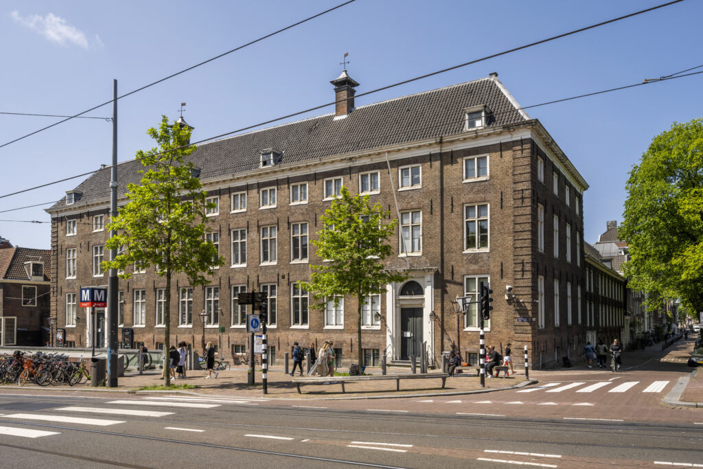 Vooraanzicht van het historische pand aan de Vijzelgracht in Amsterdam, met metro-ingang en passerende voetgangers op een zonnige dag.